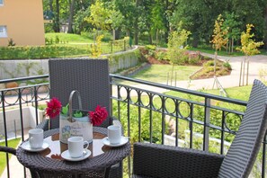 Un balcón con mobiliario de mimbre, una mesa con tazas y un jarrón de flores, y vistas a un jardín ajardinado.