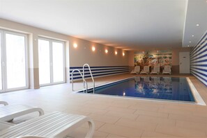 An indoor swimming pool with a tiled floor, white lounge chairs, and a mural on the wall.