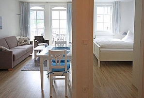 Ein Zimmer mit einer Couch, einem Esstisch mit Stühlen, einem Bett und großen Fenstern mit Vorhängen.