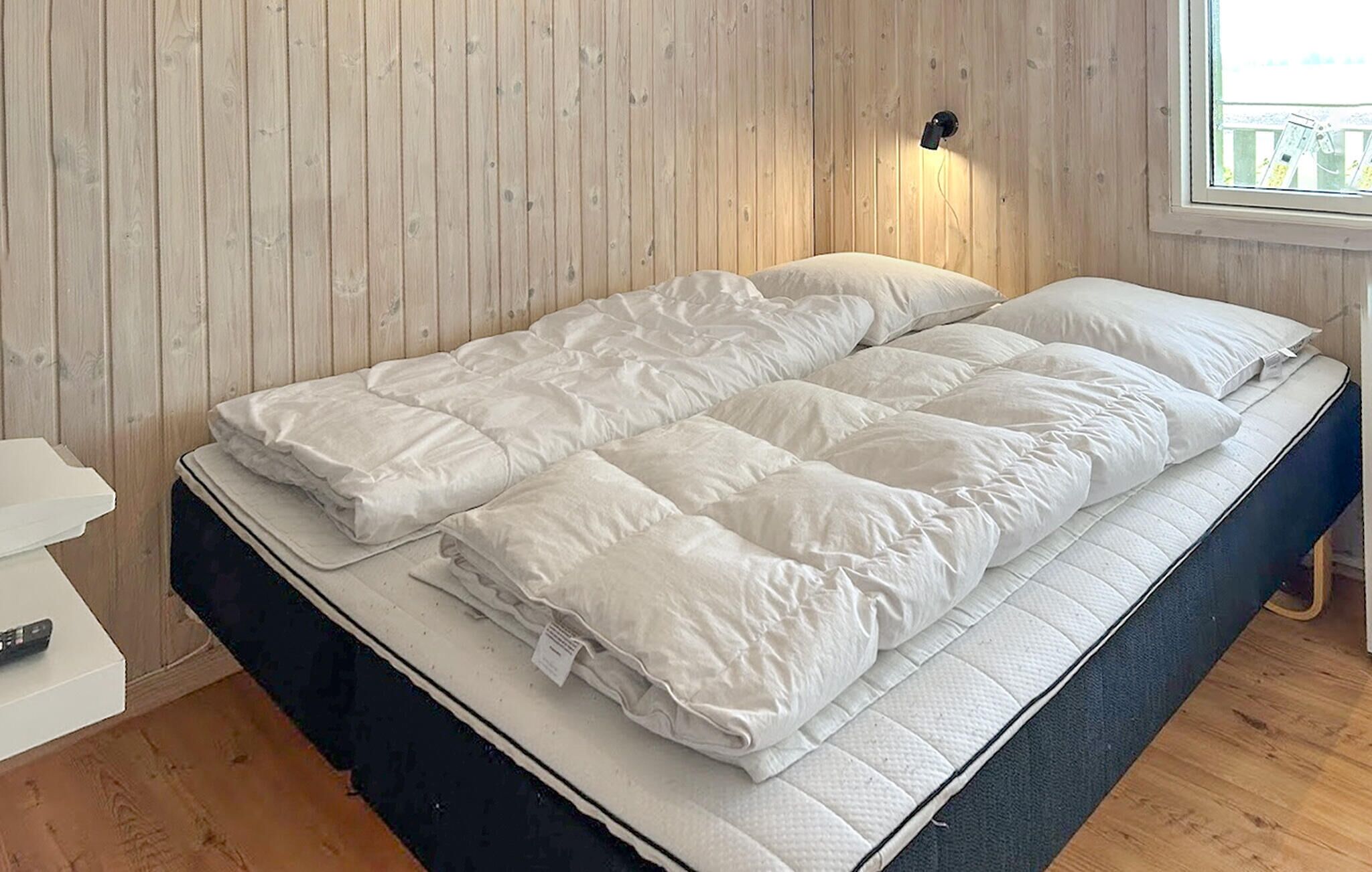 A bed with a white duvet and pillows, a bedside table with a remote control, and a window with a view of a deck.