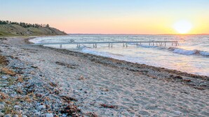 Une plage avec une jetée qui s’avance dans l’eau, un rivage rocheux et un coucher de soleil en arrière-plan.