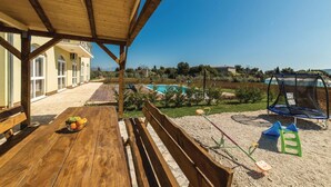 Una terraza de madera con una mesa, bancos, una piscina y un trampolín en un entorno exterior soleado.