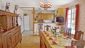 A dining area with a long table set for a meal, wooden chairs, a chandelier, and a kitchen in the background. - 6 bedroom stunning home in Pénestin (Pénestin)