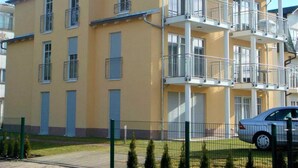 Un edificio residencial de varios pisos con balcones, un coche estacionado al frente y un patio cercado.