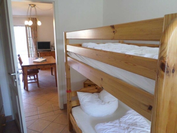 Una cama litera con ropa de cama blanca, un escritorio de madera y una silla en una habitación con suelo de baldosas.