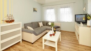 En stue med en modulsofa, et hvidt sofabord med en flaske og blomster, et fjernsyn og en skibsmodel på en hylde.