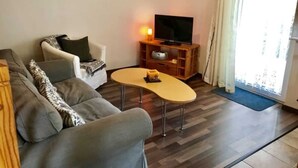 Una sala de estar con un sofá gris, una pequeña mesa redonda, un televisor sobre un mueble de madera y un ventanal con cortinas.