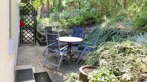 Un patio con mesa y sillas, rodeado de exuberante follaje y flores moradas.