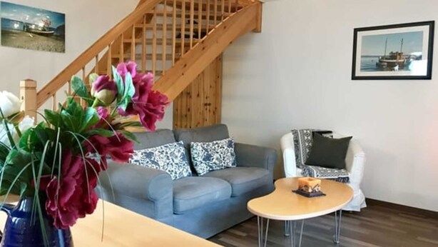 Una sala de estar con un sofá gris, una escalera de madera, una mesita redonda y un jarrón con flores.