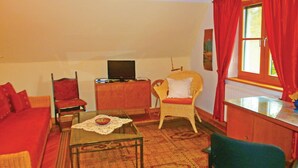 Ein Wohnzimmer mit einer Couch, einem Sessel, einem kleinen Tisch, einem Fernseher und einem Fenster mit roten Vorhängen.