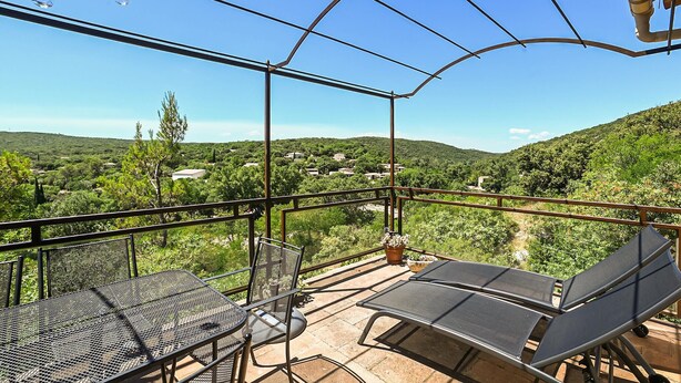 Una terraza exterior con una mesa y sillas de metal, con vistas a un paisaje verde y exuberante y un pueblo.