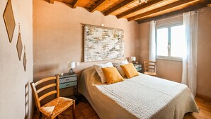 Un dormitorio con cama, una silla, mesita de noche, lámpara y ventana con cortinas.