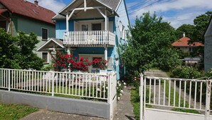 Una casa de dos pisos con balcón, rodeada por una cerca blanca y abundante vegetación.