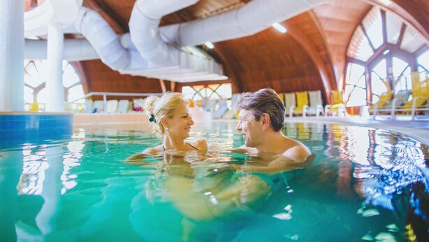 Una pareja en una piscina interior con techo de madera y ventanas amplias.