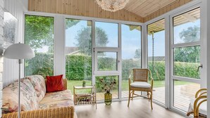 Un cuarto acogedor con un sofá de mimbre, una silla y una pequeña mesa con un jarrón. Amplias ventanas ofrecen vistas a un jardín.