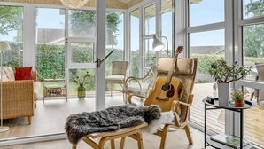 Una habitación con un gran ventanal que da a un jardín, un sofá de mimbre, una silla de madera con una guitarra, una mesita con una lámpara y un banco con un plaid de piel.