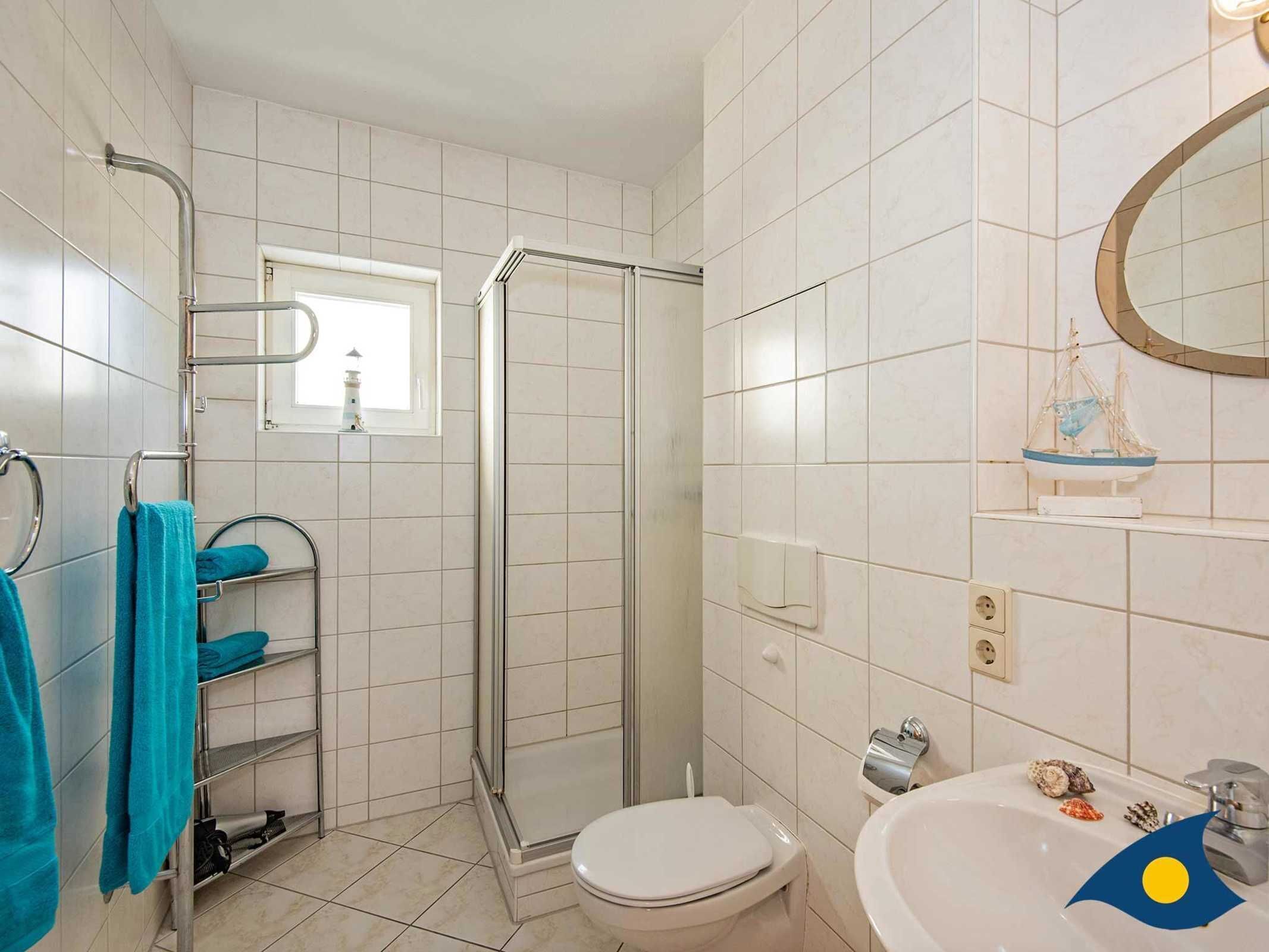 Ein Badezimmer mit Duschkabine aus Glas, Toilette, Waschbecken mit dekorativen Muscheln und einem Spiegel mit Schiffsmotiv.