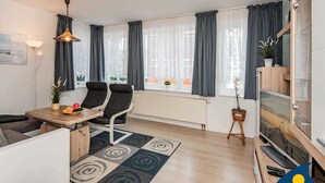A modern living room with a sofa, two black chairs, a dining table with fruit, a lamp, and a TV on a wooden stand. - Blaue Welle Whg. 11 Marius // - BW 11 (Heringsdorf)