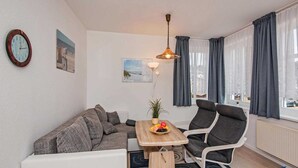 A modern living room with a sofa, a dining table with fruit, a chair, a lamp, and a view of the outdoors through large windows. - Blaue Welle Whg. 11 Marius // - BW 11 (Heringsdorf)