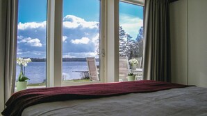 Un dormitorio con un ventanal grande que da a un lago y árboles, una cama con una colcha roja y un jarrón con flores blancas en el alféizar.