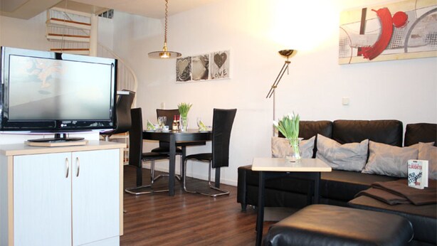 A modern living room with a flat-screen TV, a dining area with chairs and a table, a sofa, and a small table with a lamp. - (50) 3-room apartment - apartment house Mecklenburg (Kühlungsborn)