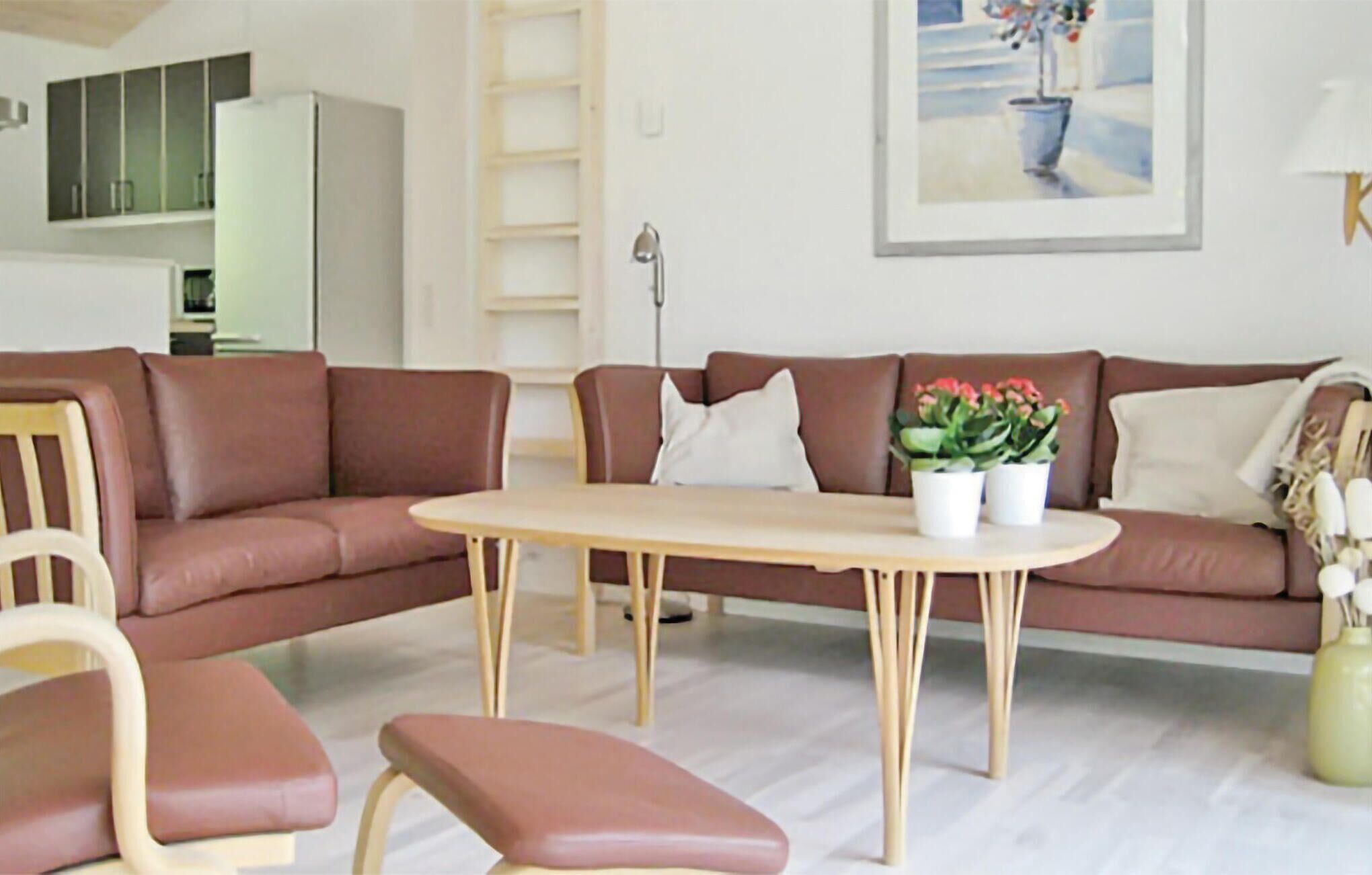 Sala de estar com um sofá de couro marrom, uma mesa de centro de madeira e uma poltrona branca. Há um quadro na parede e um abajur em uma mesinha lateral.