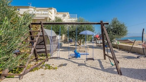 A playground with swings and a slide, surrounded by gravel and greenery, with buildings in the background. - Stunning apartment in Vir with house sea view (Vir)