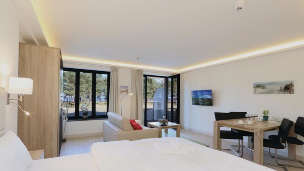 A modern hotel room with a large bed, a desk, and a dining table. There are large windows offering a view of the outdoors.