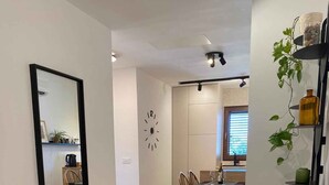Una sala de estar moderna con un sofá gris oscuro, una mesa de centro redonda de madera y un espejo grande en la pared. Se observa un comedor con una mesa y sillas al fondo.