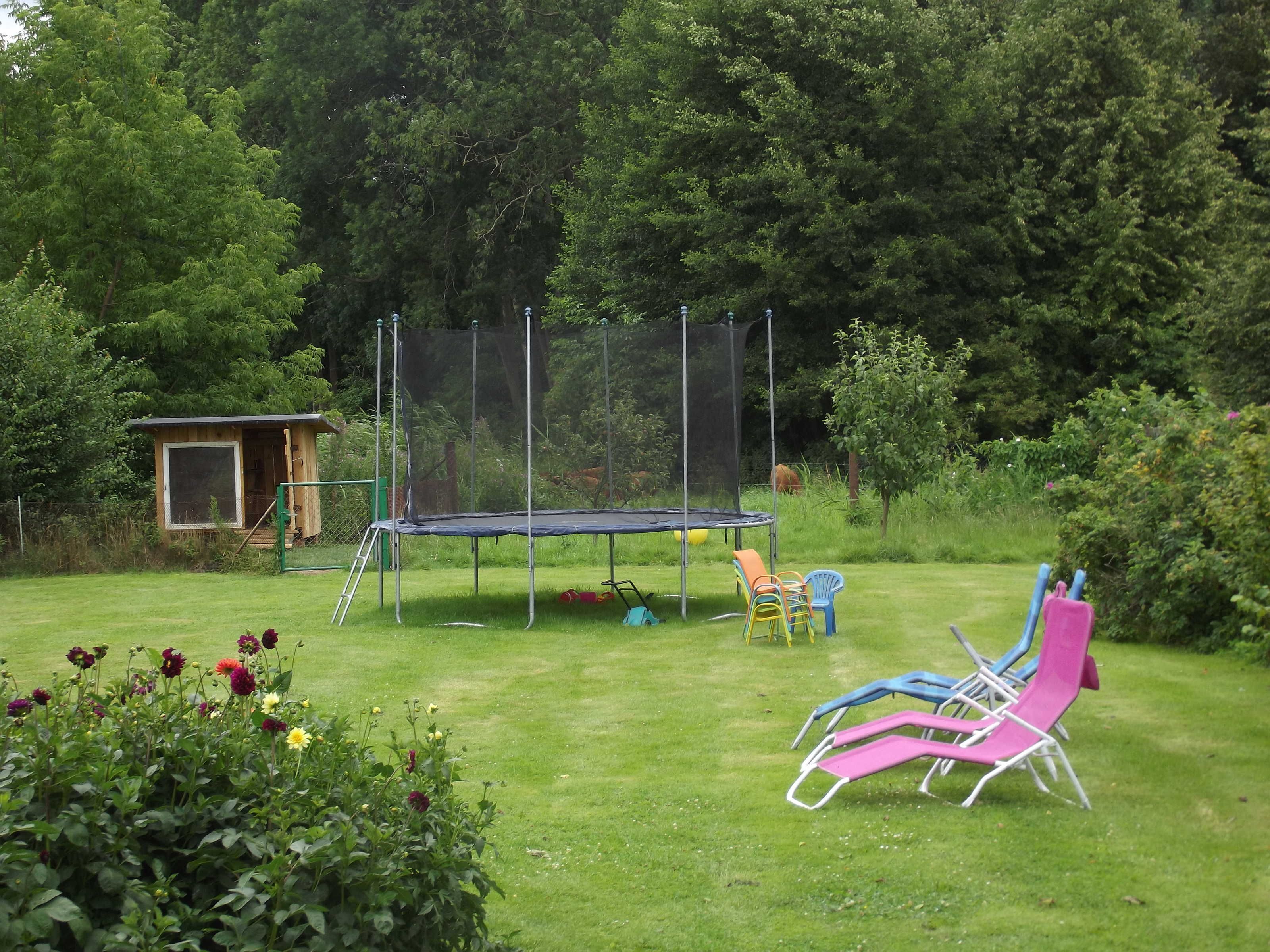 Un trampolín en un patio trasero con césped, una pequeña estructura de madera y sillas coloridas.