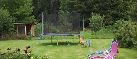 Un trampolín en un patio trasero con césped, una pequeña estructura de madera y sillas coloridas.