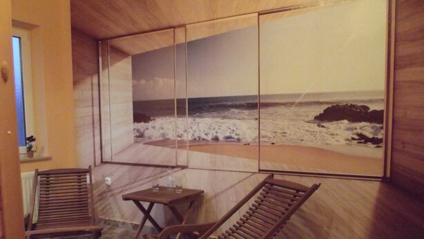 Une pièce décorée d’une grande fresque représentant une plage, avec deux fauteuils à bascule en bois, une petite table et une fenêtre agrémentée d’un pot de fleurs.