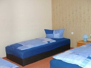 Een kamer met twee éénpersoonsbedden, elk met blauw beddengoed en kussens, een houten wand, een klein nachtkastje met een lamp en een eenvoudig vloerkleed.