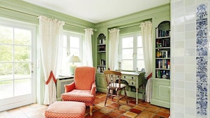 Une pièce avec un mur vert, un fauteuil à motifs rouges, une chaise en bois, un bureau et une bibliothèque.
