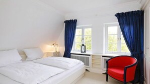 Une chambre à coucher avec un lit, deux fenêtres avec des rideaux, un radiateur, une table de chevet et un fauteuil rouge.