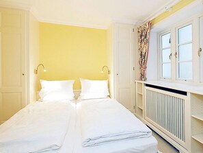 Uma cama de casal com roupa de cama branca em um quarto com paredes amarelas, um radiador e uma janela com cortinas.
