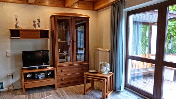 A living room with a wooden cabinet, a television, a small table, and a large window. - Heisch: Cozy vacation home with terrace and beach chair - Ferienhaus Heisch (Schafflund)