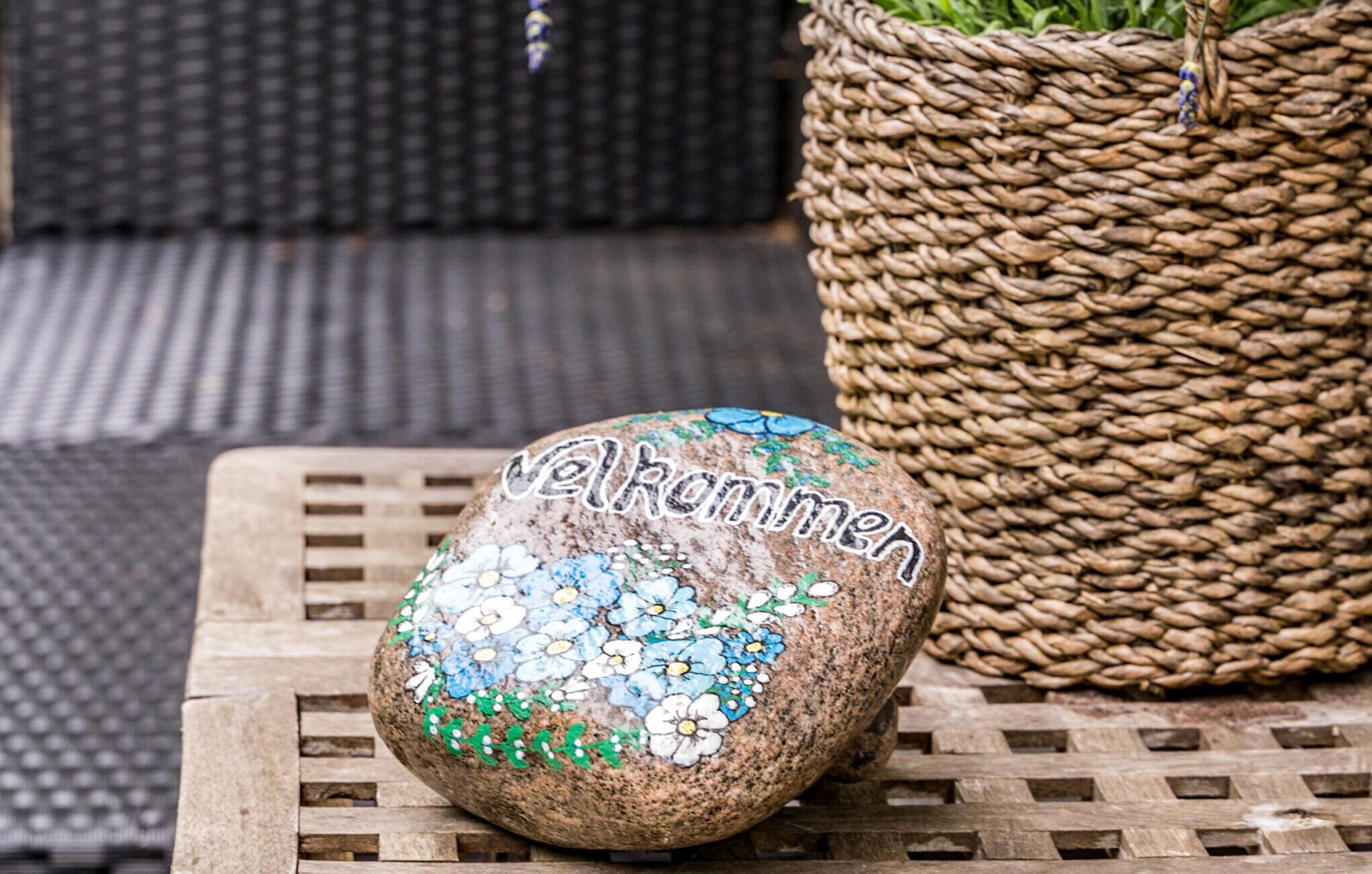 A painted rock with "Valhommers" on it, placed on a wooden table next to a woven basket.