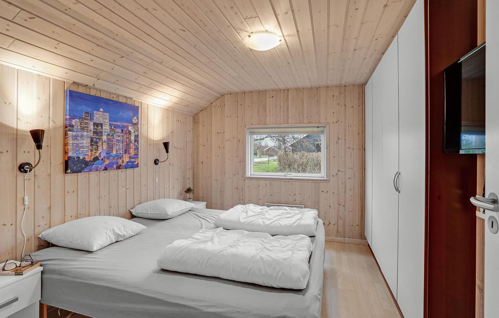 Une chambre avec des murs recouverts de panneaux en bois, un lit avec une literie blanche, un tableau représentant un paysage urbain, une fenêtre offrant une vue et un téléviseur fixé au mur.