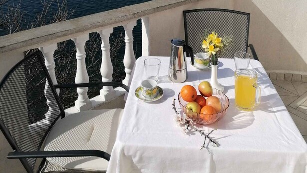 A table set with a white tablecloth, a bowl of fruit, a pitcher of juice, and a cup on a balcony overlooking a body of water. - Holiday apartment near the sea (Barić Draga)