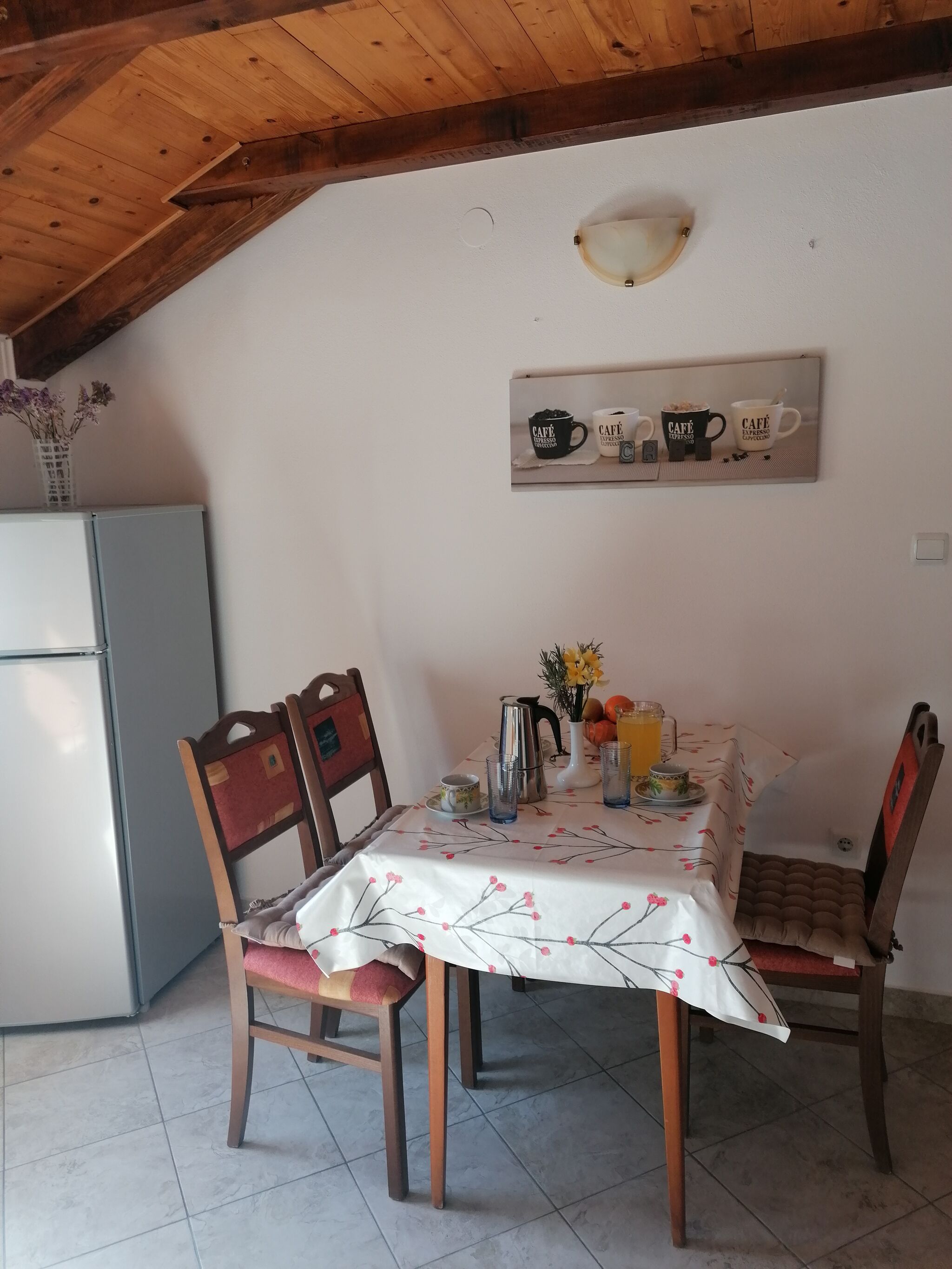 A dining area with a table set for two, chairs, a refrigerator, and a wall-mounted light fixture.