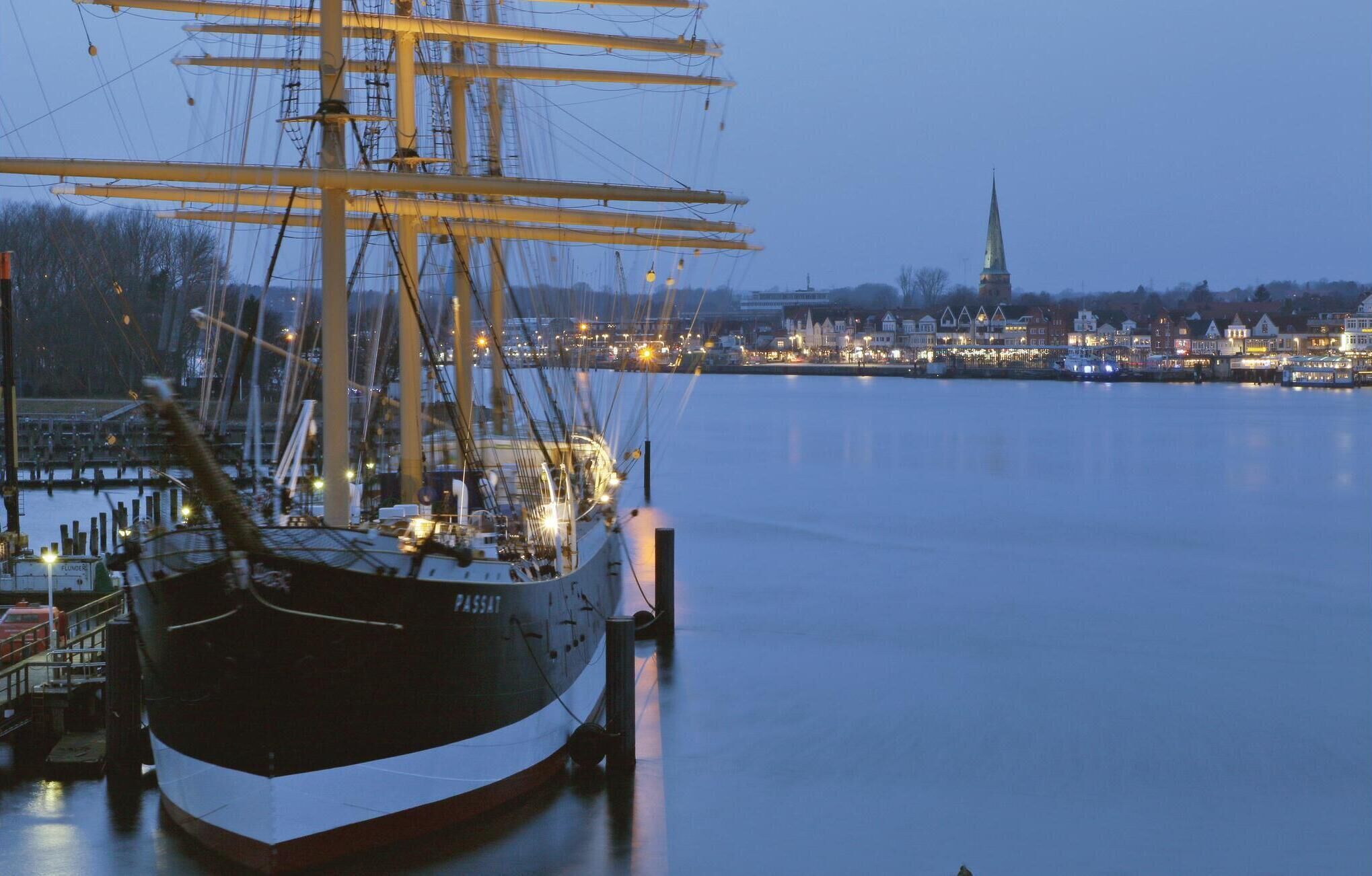 A large sailing ship docked at a harbor with a cityscape in the background.
