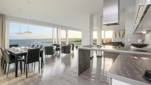 Een moderne keuken met een groot eiland, een eethoek met uitzicht op zee, en een ruime woonkamer met hoge plafonds en grote ramen.