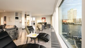 Een moderne hotelkamer met een groot raam met uitzicht op een stadslandschap en water. Het heeft een bank, een eethoek en een kitchenette.
