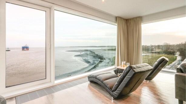 Een moderne kamer met een groot raam met uitzicht op een strand en een schip, een gebogen bank en een kleine tafel met een fles en een glas.