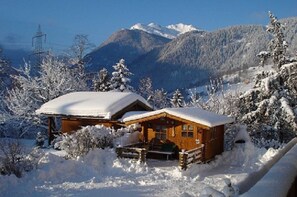A wooden cabin with a snow-covered roof, surrounded by snow-covered trees and mountains in the background.