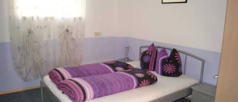 A single bed with a metal frame, a bedside table, a window with sheer curtains, and a framed picture on the wall.