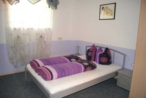 A single bed with a metal frame, a bedside table, a window with sheer curtains, and a framed picture on the wall. - Apartment Ursula - Apartment 1 (Grins)