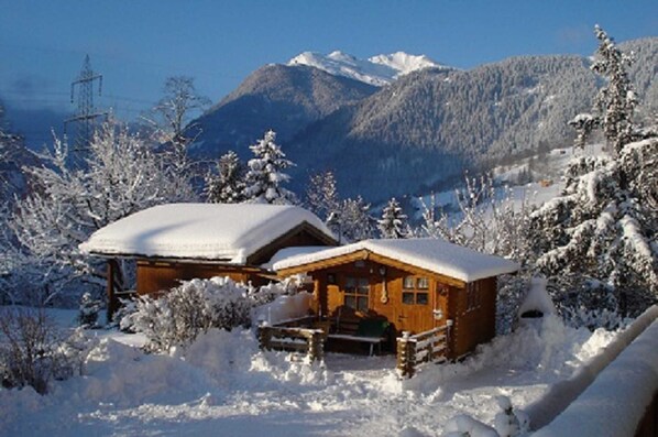 A wooden cabin with a snow-covered roof, surrounded by snow-covered trees and mountains in the background. - Apartment Ursula - Apartment 1 (Grins)