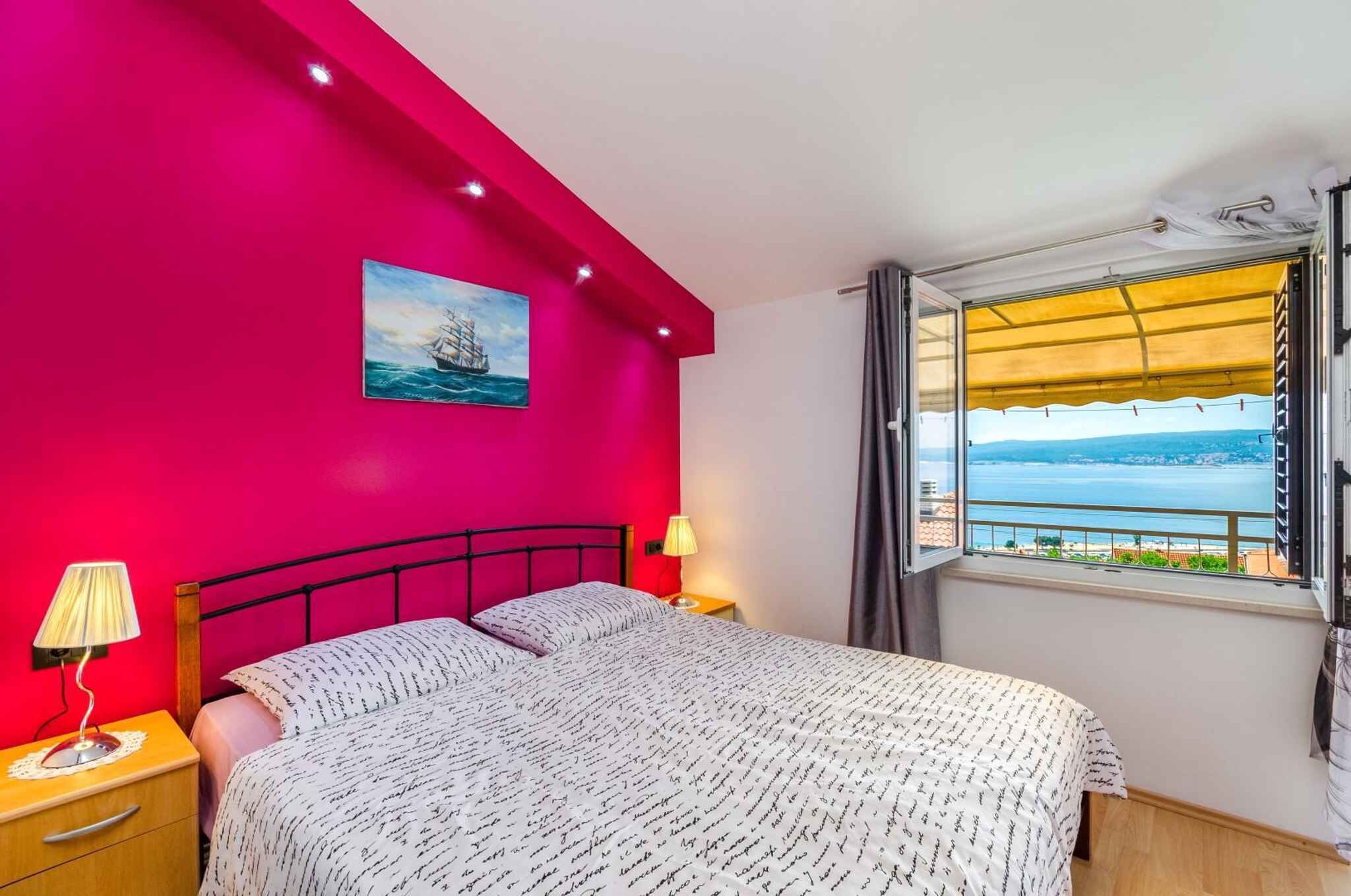 A bedroom with a bed, a nightstand, a lamp, a painting, and a window with a view of the sea.